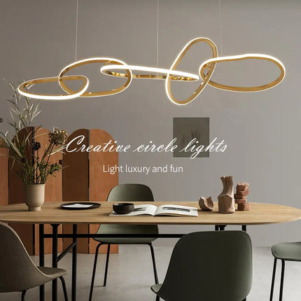 A gold pendant light chandelier with a modern design, hanging over a wooden dining table in a minimalist setting, surrounded by black chairs and decorative objects.