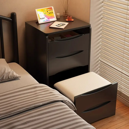 A dark brown nightstand with two drawers and a matching ottoman in a bedroom setting next to a bed with striped bedding.