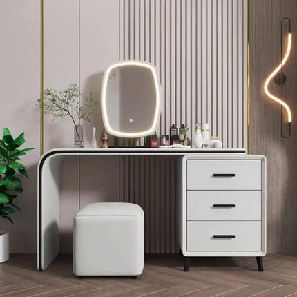 A modern makeup vanity table set in a styled room, featuring a white desk with three drawers, a matching white stool, and an oval LED-backlit mirror. The background consists of a slatted wood wall panel and a warm wood floor.