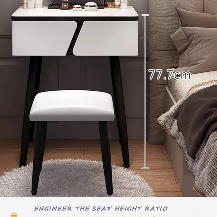 A white vanity desk with two drawers and black legs, paired with a matching white upholstered stool. The scene is set in a bedroom next to a bed, with a height measurement overlay reading 77.