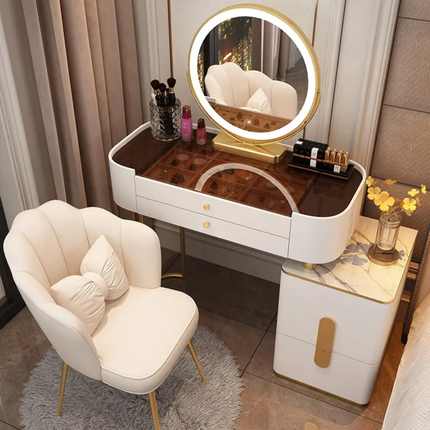 A modern vanity table set in a bedroom, featuring a white and gold makeup desk with a built-in oval LED mirror, a matching white armchair, and a marble-top side cabinet.
