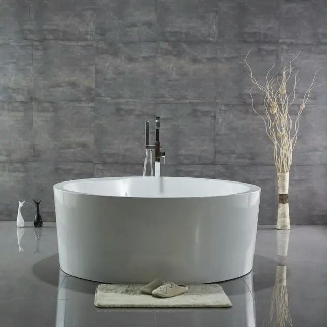 A round freestanding bathtub in glossy white, set against a gray stone-look tiled wall with a matching gray floor. A tall chrome faucet is mounted behind the tub, and a small folded bath mat with a pair of beige slippers rests in front.