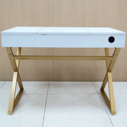 A modern writing desk with a white rectangular top, a single drawer, and a gold-finished metal X-shaped base, set against a light wood paneled wall and white tiled floor.