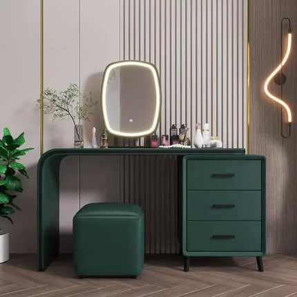 A modern vanity desk set in dark green, featuring a three-drawer cabinet, a matching green stool, and an oval LED-backlit mirror. The set is styled in a contemporary bathroom or dressing area with a slatted wood wall panel, a glowing wavy wall sconce, and a potted plant.