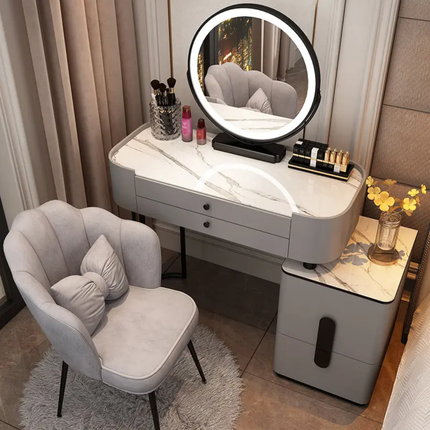 A modern vanity desk setup in a bedroom or dressing area, featuring a white marble-patterned makeup table with an illuminated oval mirror, a matching gray upholstered chair with pillows, and a side cabinet with a marble top. The room has beige curtains, tiled flooring, and neutral decor.