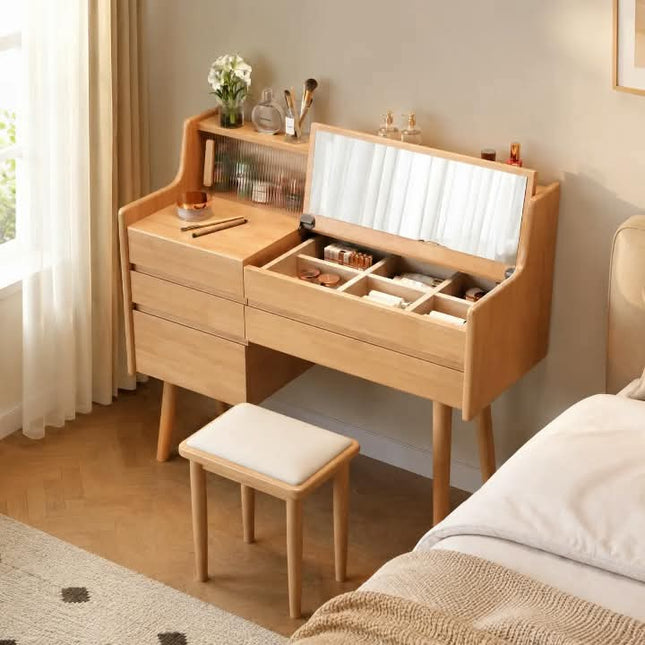 A light natural wood vanity desk with an open top mirror and storage compartments, shown in a bedroom setting next to a bed with a matching wooden stool.