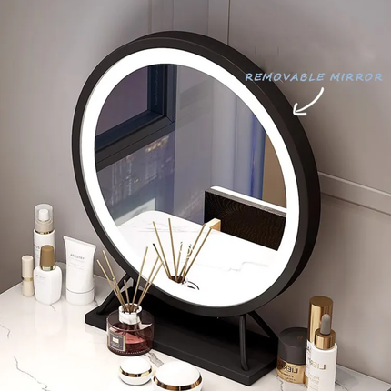 A round LED vanity mirror with a black frame and stand, shown on a white marble countertop with cosmetics and a reed diffuser. The mirror has a white ring light around the edge, and a blue text overlay reads 'REMOVABLE MIRROR' with an arrow pointing to the frame.