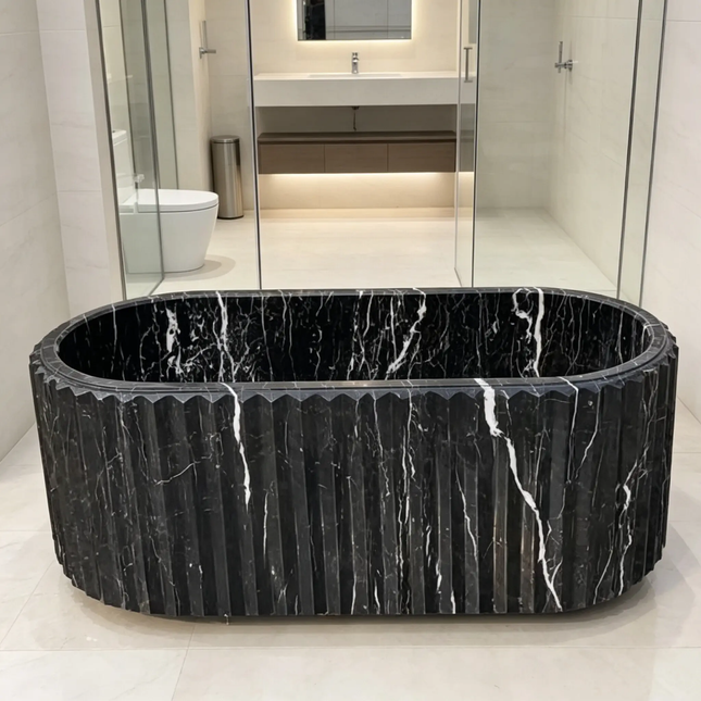 A freestanding oval bathtub made of black marble with white veining, featuring a fluted or ribbed exterior, set in a modern bathroom with light beige tiles and glass walls.