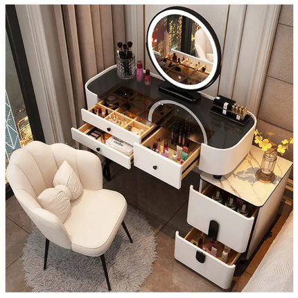 A modern makeup vanity table set in a bedroom, featuring a white and black frame, an illuminated oval mirror, multiple drawers, a marble-topped side table, and a matching cream-colored accent chair.