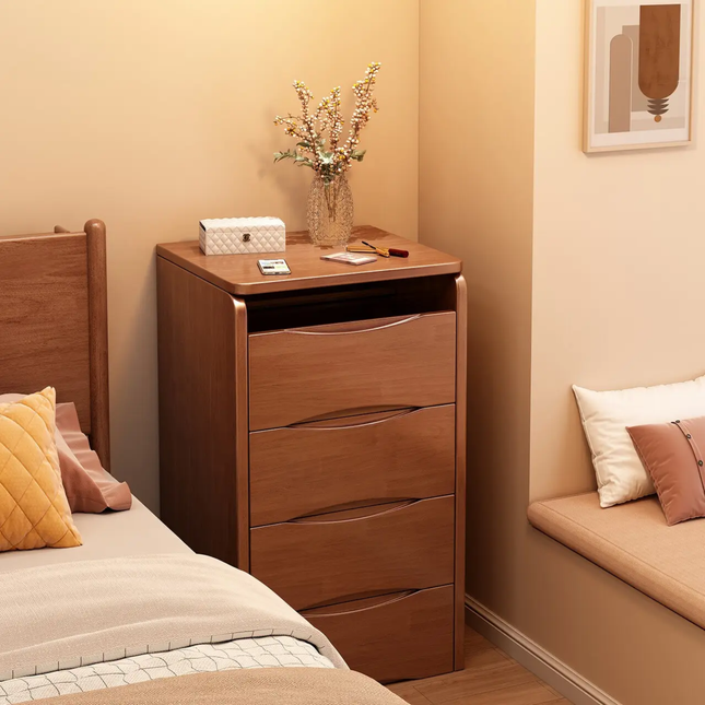 A four-drawer wooden nightstand or storage cabinet in a warm medium brown finish, styled in a bedroom next to a bed with beige bedding and pillows. The cabinet has a top surface holding a glass vase with dried flowers, a small white box, and a small card holder.