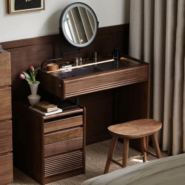 A dark brown wooden vanity table set in a bedroom, featuring a built-in round mirror, a side drawer with three drawers, a small vase with pink tulips, and a matching wooden stool.