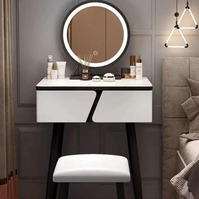 A modern makeup vanity table with an integrated oval mirror, white front panels, black legs, and a matching white stool, styled in a bedroom setting.