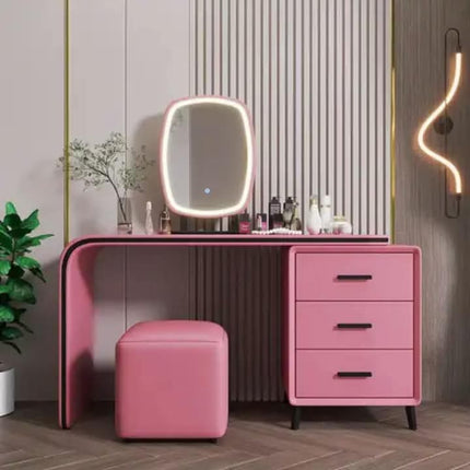 A pink vanity desk set in a modern bathroom or dressing area, featuring a three-drawer cabinet, a matching pink stool, and a built-in oval mirror with a glowing LED light frame. The furniture is styled against a slatted wood-paneled wall with a decorative plant and a wall sconce.