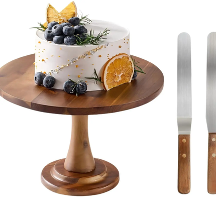 A round wooden cake stand holding a decorated white cake with blueberries, rosemary, a dried orange slice, and gold leaf accents, shown on a white background with two spatula tools at the right.