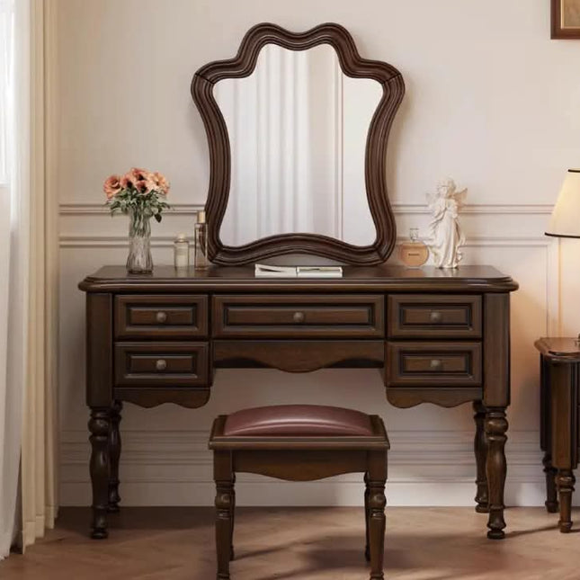 A dark brown wooden vanity table with an attached scalloped mirror and matching stool, styled in a classic bedroom or dressing room setting with a vase of flowers and a lamp.