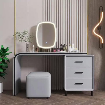 A modern gray vanity desk with an integrated oval LED-lit mirror, three drawers on the right, and a matching gray stool, styled in a contemporary bedroom or dressing area with wood slat wall panels and warm ambient lighting.