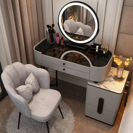 A modern makeup vanity table set in a bedroom, featuring a light gray frame, a built-in round LED mirror, a matching side cabinet, and a curved gray velvet accent chair with pillows.