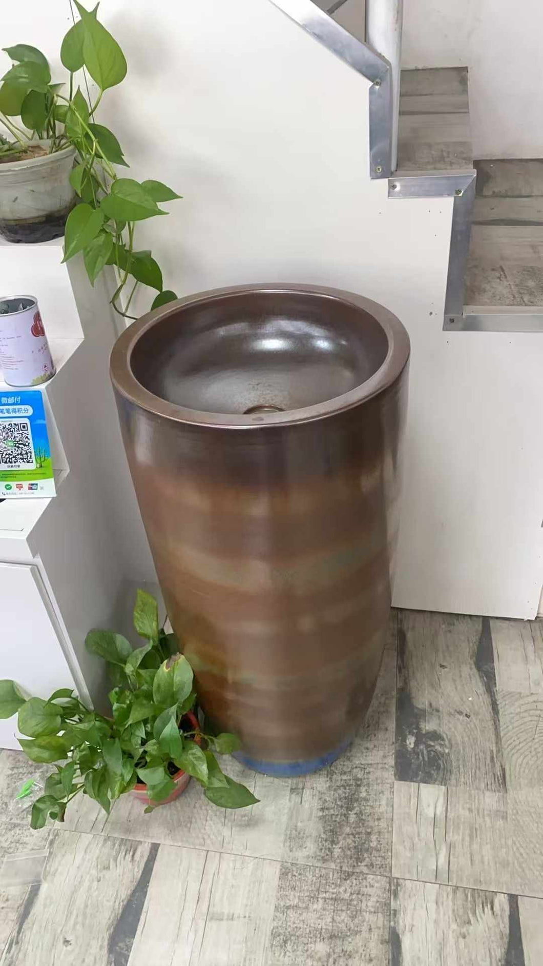 Tall Cylindrical Freestanding Wash Basin with Metallic Bronze Finish, Made of High-Quality Ceramic or Stone, Featuring a Smooth, Polished Interior and a Minimalist Design, Complemented by Greenery in a Modern Indoor