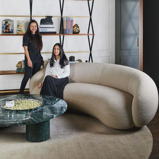 Two women posing in a modern living room with a curved beige sofa and marble coffee table.