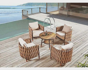 Outdoor patio set with wicker chairs and table on a wooden deck by a pool and ocean view.