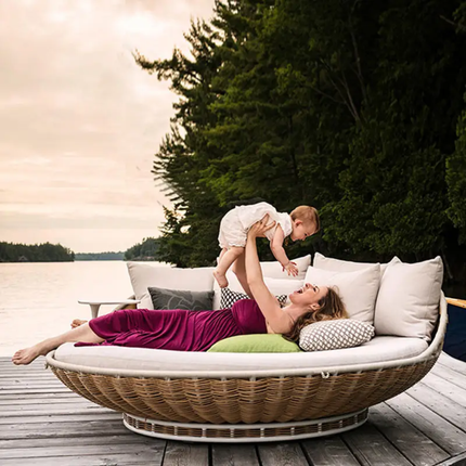 Woman in a pink dress lying on a circular outdoor sofa by a lake, holding a baby.