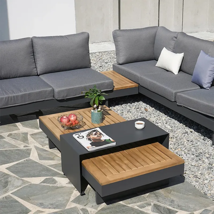 Outdoor patio set with gray sectional sofa, wooden coffee table, and decorative items on a stone patio.