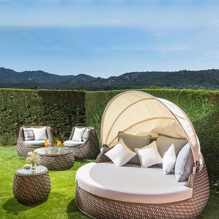 Outdoor patio set with a round daybed, chairs, and tables on a grassy area with a scenic background.