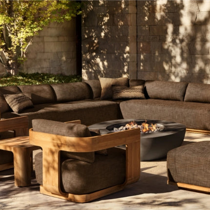 Outdoor patio furniture set with brown sectional sofa, wooden tables, and ottomans in a sunlit area.