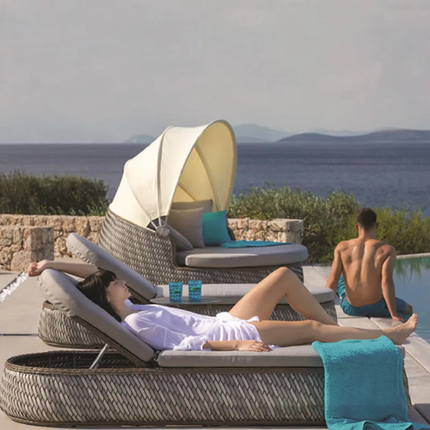 Two people relaxing on wicker sun loungers by a pool with a scenic view of mountains.