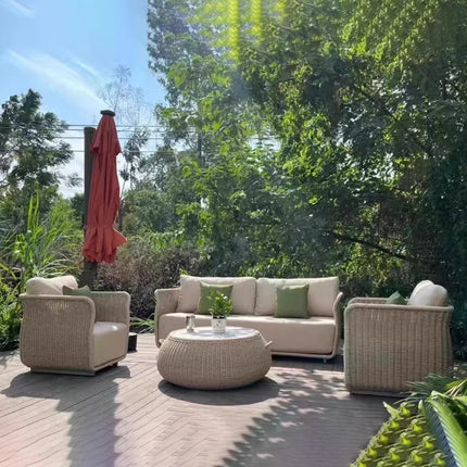Outdoor patio set with beige sofa, chairs, and ottoman on a wooden deck with greenery in the background.