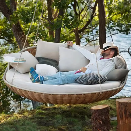 Man relaxing on a hanging outdoor sofa by a body of water with trees in the background.