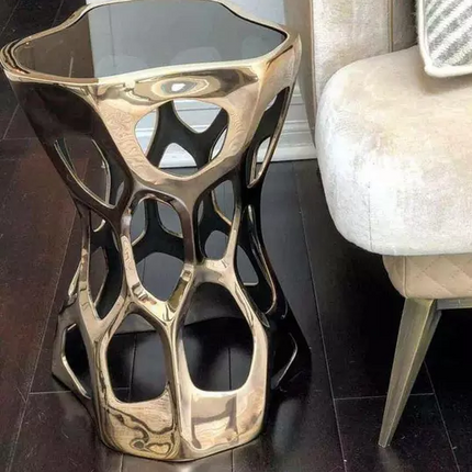 A stainless steel side coffee table with a geometric sculpture design, featuring a mirror polished finish, placed on a hardwood floor next to a beige sofa cushion.