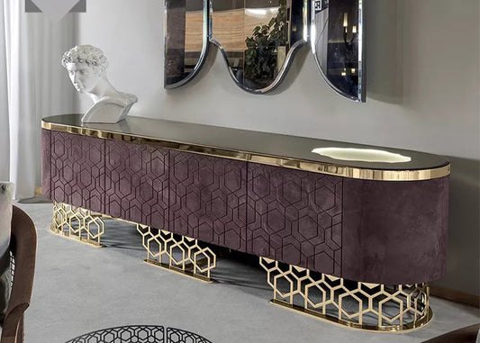 A modern TV stand with a purple and gold color scheme, featuring a flat-screen TV on top, set against a reflective wall with a large mirror behind it. The stand has a geometric pattern on the legs and a drawer for storage.